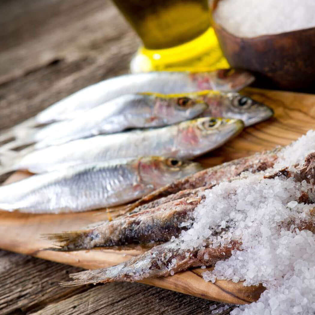 Fresh and salted anchovies on a wooden board with sea salt, representing Acciughe Intere Salate 1 kg from the Mediterranean, processed in Sicily.